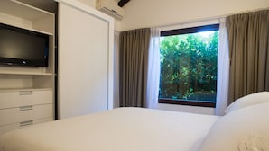 A hotel room with a bed, a TV on a stand, a window with curtains, and a view of greenery outside.