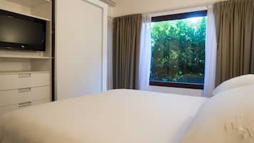 Habitación de hotel con cama, televisor sobre un mueble, ventana con cortinas y vista a zonas verdes.