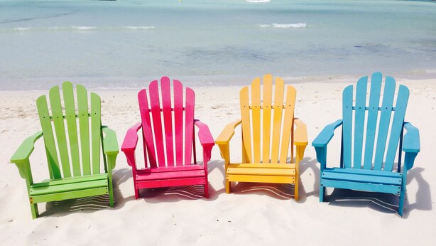 Four colorful Adirondack chairs lined up on a sandy beach with the ocean in the background. - Tu Casita (Noord)