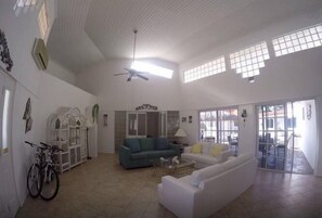 A spacious living room with a high ceiling, a ceiling fan, and large windows allowing natural light. - Tu Casita (Noord)