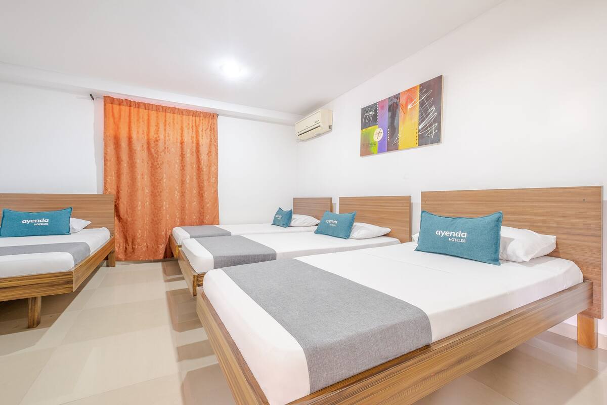 Une chambre d’hôtel avec quatre lits, chacun doté d’une tête de lit en bois et d’une couverture grise. Un climatiseur est fixé au mur, et une peinture abstraite aux couleurs vives est accrochée au-dessus des lits.