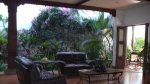 A spacious living area with a large sofa, a coffee table, and a dining table. There is a view of lush greenery outside. - Hotel Nicarao Inn (Rivas)