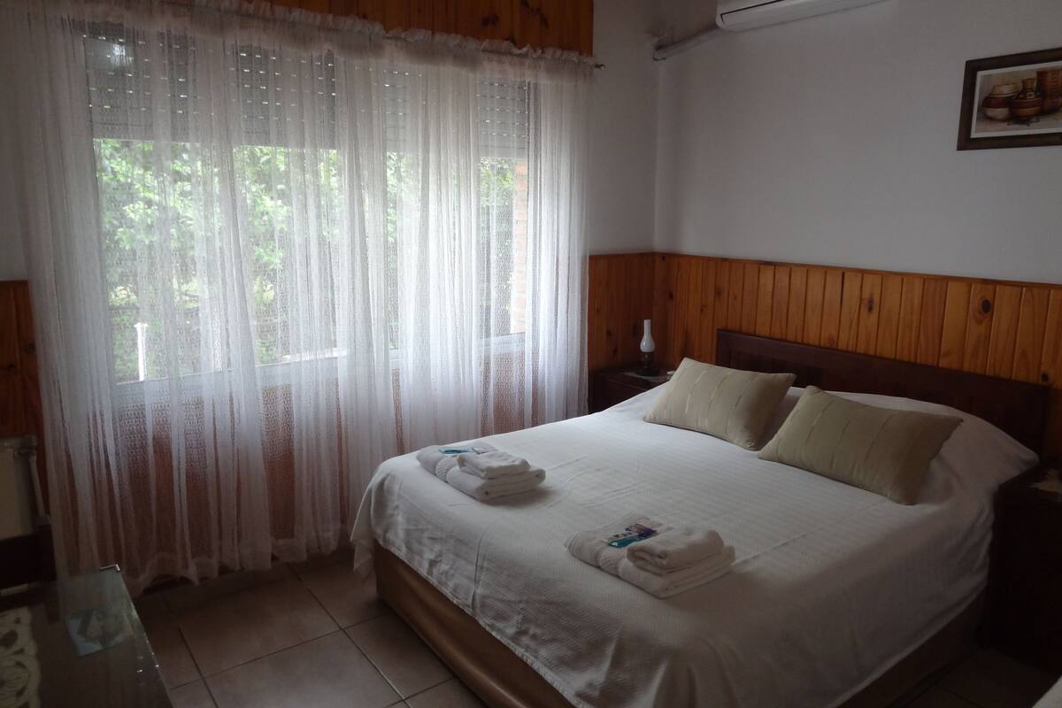 Un dormitorio con cabecera de madera, cama con ropa blanca y ventana con cortinas de gasa.