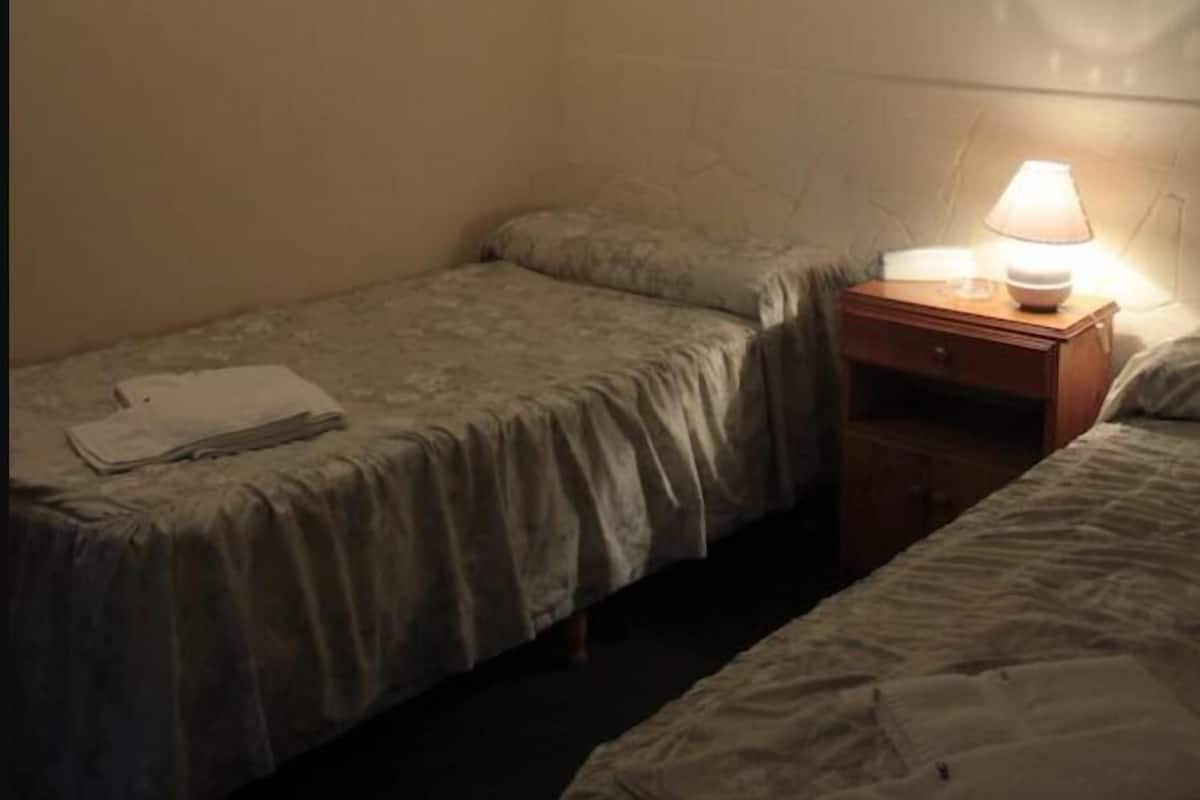 A small, dimly lit room with two single beds, a nightstand with a lamp, and a folded piece of paper on one bed.