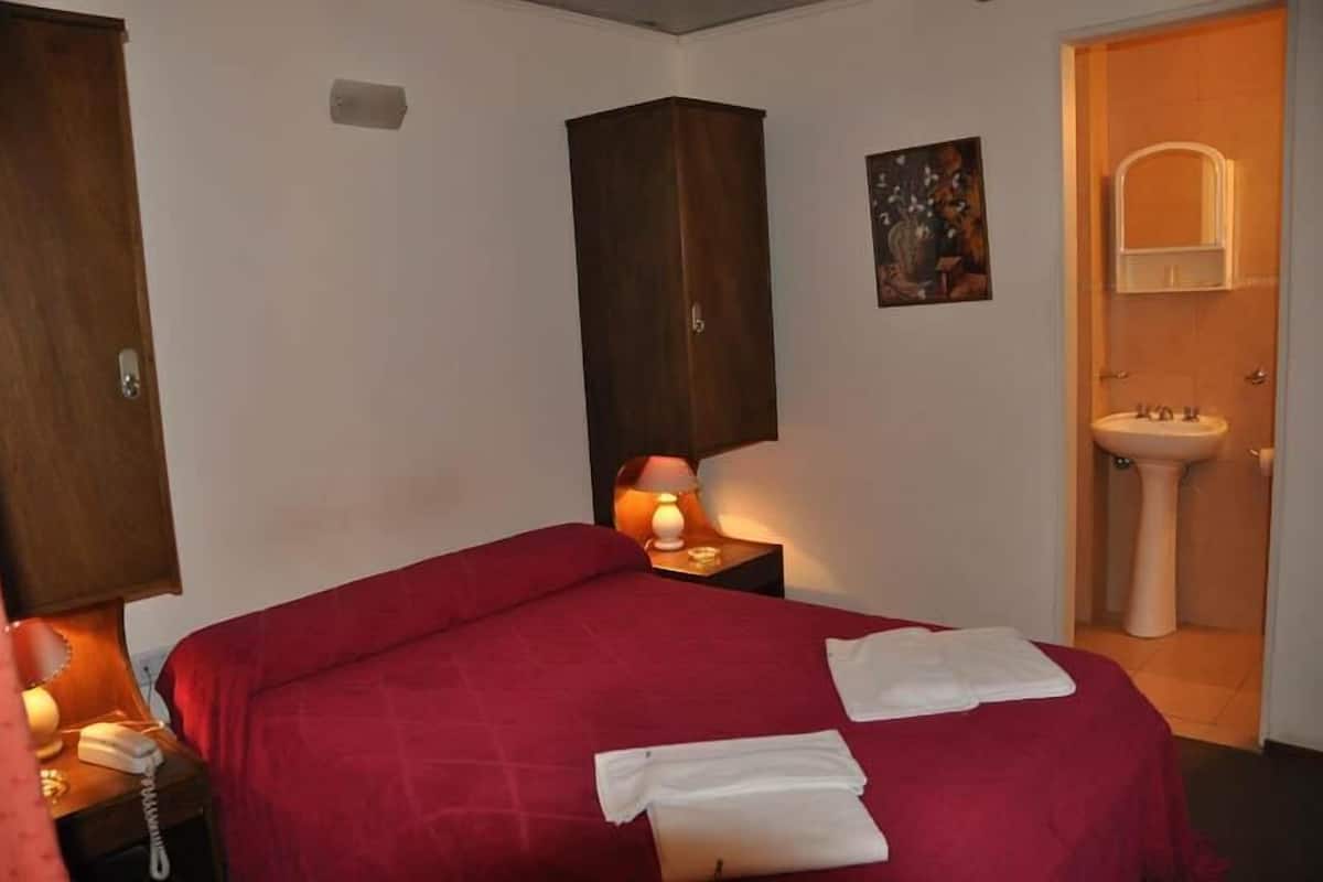 A hotel room with a bed, a bedside lamp, a telephone, a painting on the wall, and a bathroom with a sink and mirror.