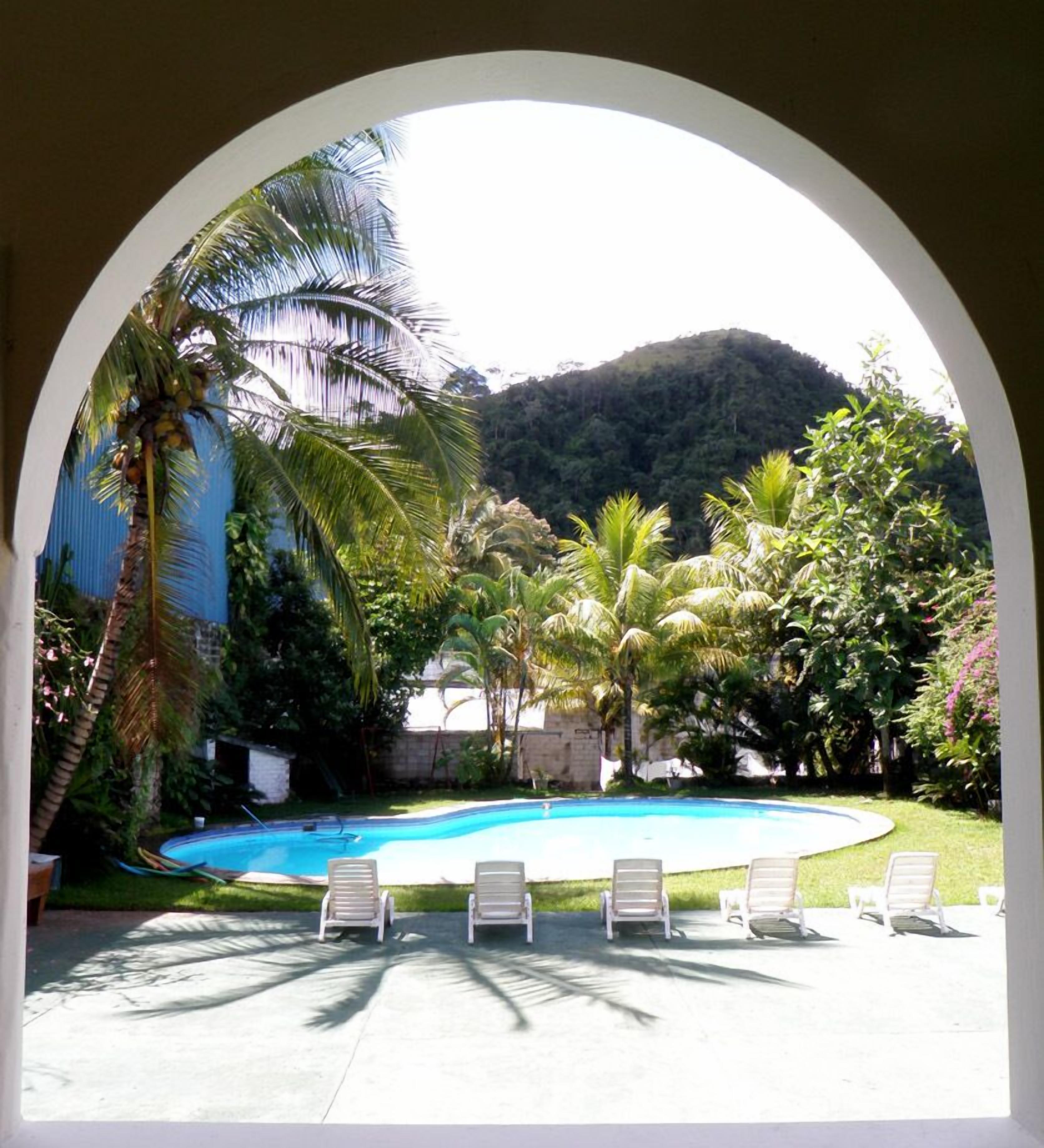 A swimming pool with lounge chairs, surrounded by palm trees and a mountainous backdrop.