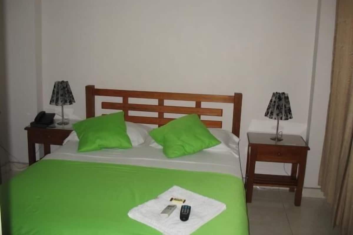 A neatly made bed with green pillows, a bedside table with a lamp, and a remote control on the bed.