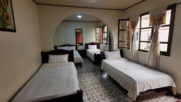 Quarto de hotel com quatro camas individuais dispostas em linha, piso ladrilhado e uma grande janela em arco.