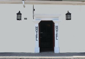 Entrada de un hotel con fachada blanca, puerta de acceso oscura y dos lámparas negras de estilo farol a cada lado.
