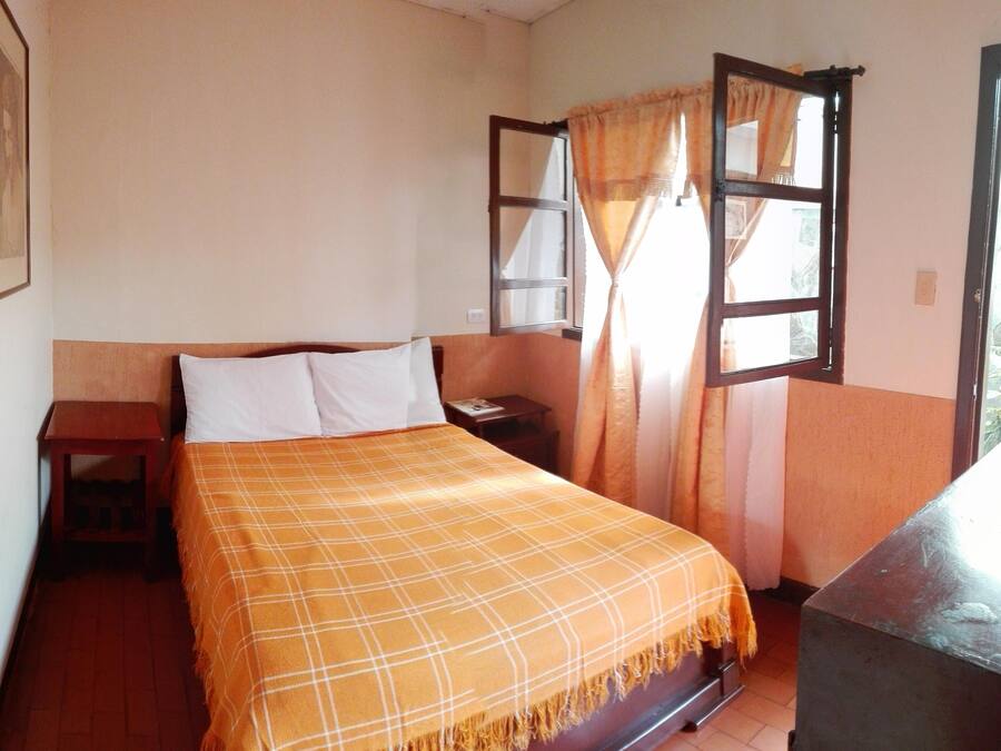 A bedroom with a bed, a nightstand, a window with curtains, and a wall-mounted shelf.