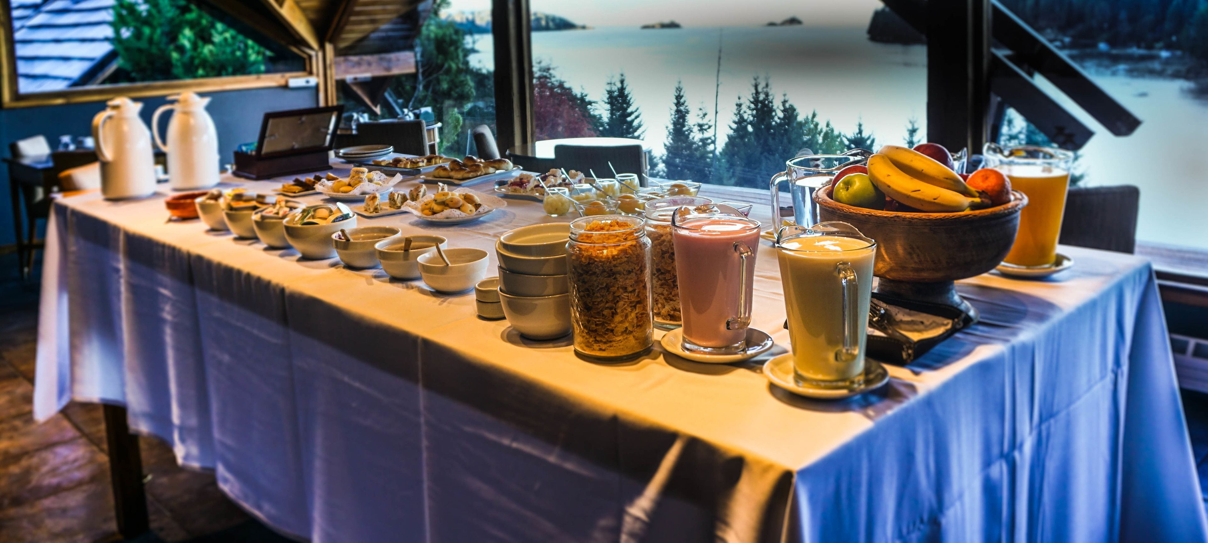 Une table de buffet proposant une variété de plats et de boissons, le tout installé dans un cadre pittoresque avec vue sur des arbres et un lac.