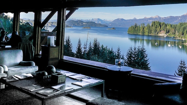 Habitación acogedora con un ventanal grande que da a un lago y montañas. Hay un sofá y una pequeña mesa con papeles.
