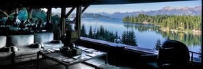 A cozy room with a large window overlooking a lake and mountains. There is a sofa and a small table with papers. - Marinas Alto Manzano (Villa La Angostura)