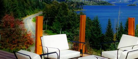 A deck with two white cushioned chairs and a small table, overlooking a lake and forested area.