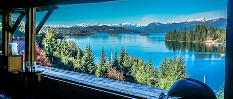 Una habitación con un ventanal grande que da a un lago, rodeada de árboles y montañas.