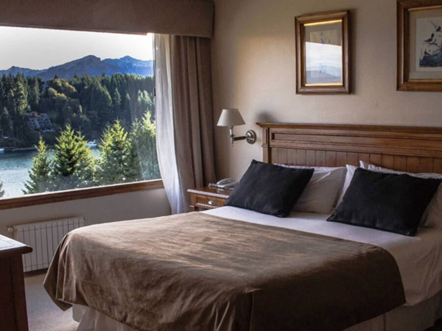 Una habitación de hotel con una cama, dos mesitas de noche, una lámpara y vistas a un lago y montañas.