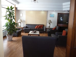 Una sala de estar con un sofá, sillones, una mesa de centro, un televisor, una planta en maceta y un ventanal con persianas.