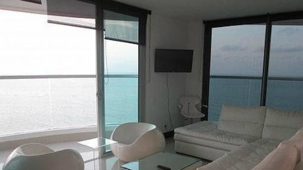 Uma sala de estar moderna com um sofá branco, uma poltrona branca e uma mesa de centro de vidro. Há uma TV de tela plana na parede e uma grande janela com vista para o mar.