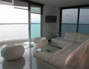 A modern living room with a white sofa, a white armchair, and a glass coffee table. There is a flat-screen TV mounted on the wall and a large window offering a view of the sea. - Palmetto Eliptic Apartamentos (Cartagena de Indias)