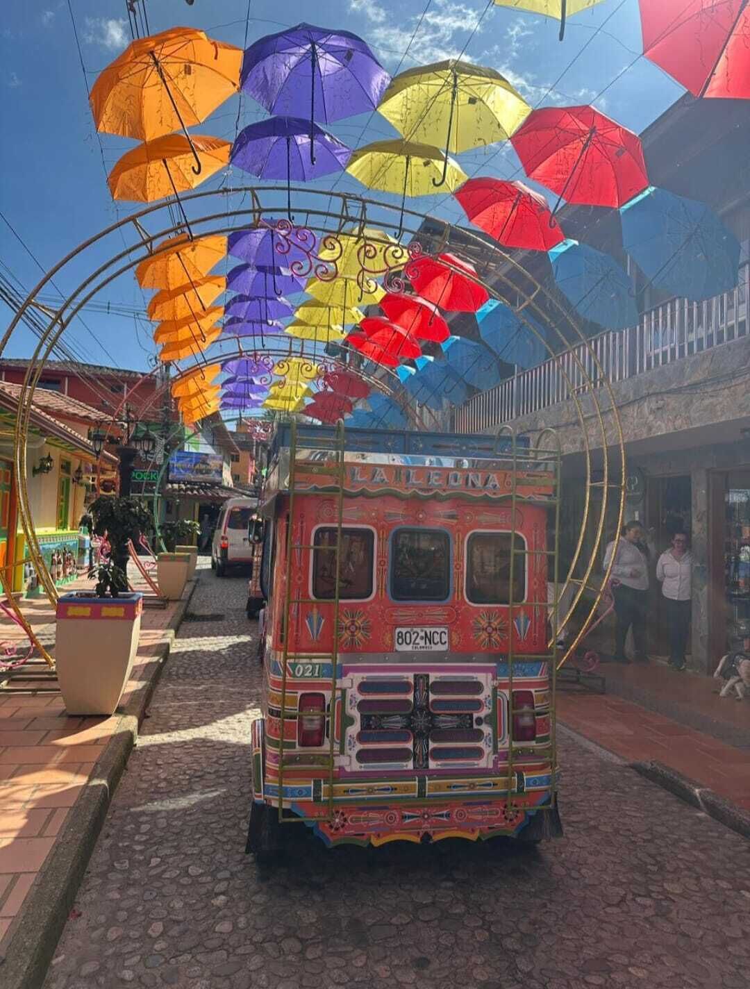 Un autobús colorido con un diseño llamativo estacionado en una calle empedrada, rodeado por un toldo de sombrillas.