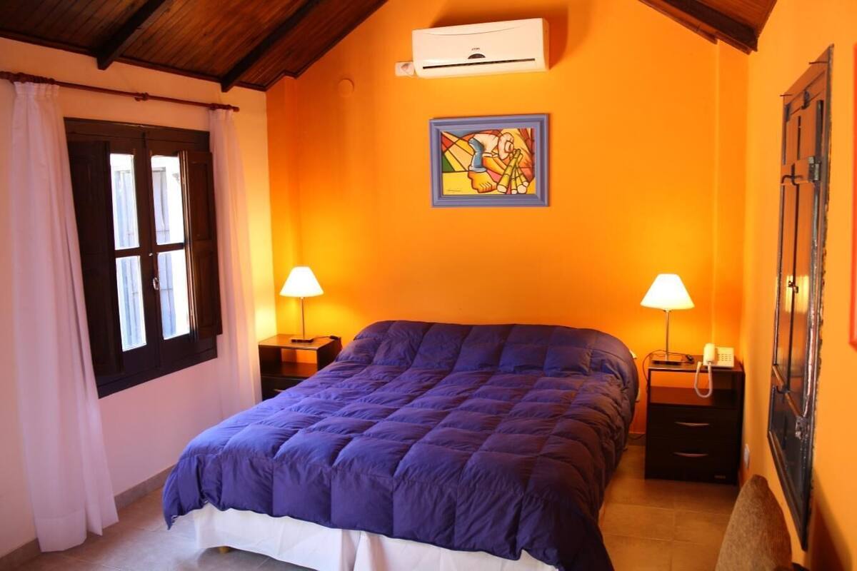 Quarto com uma cama grande, duas mesas de cabeceira com luminárias, ar-condicionado fixado na parede e uma janela com cortinas.