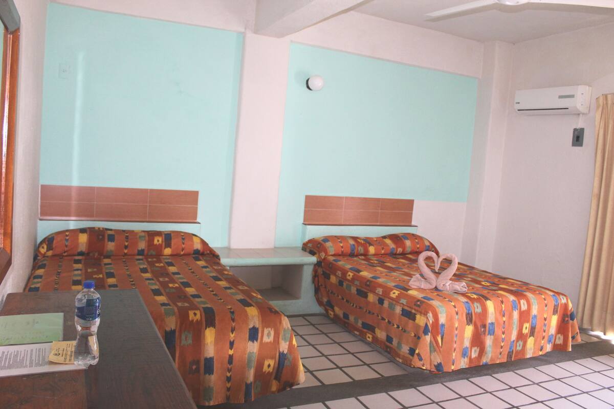 A hotel room with two beds, a small table, a water bottle, and an air conditioning unit.