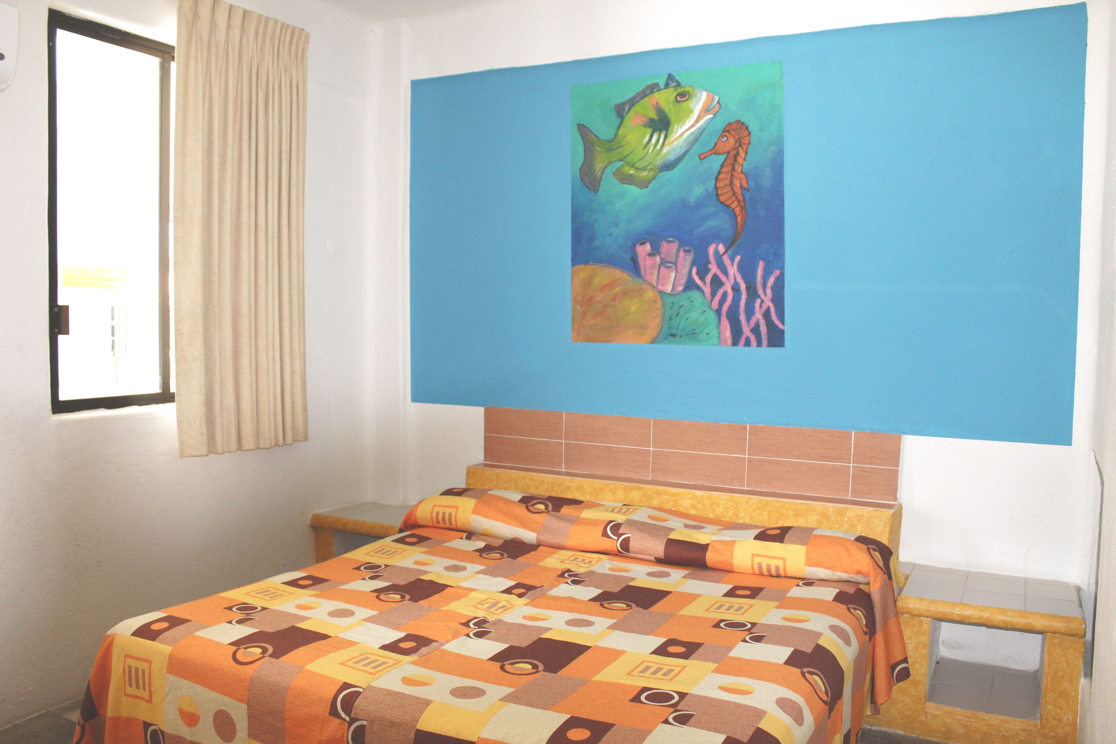 Habitación de hotel con cama, ventana con cortinas y un cuadro de un pez y un caballito de mar en la pared.