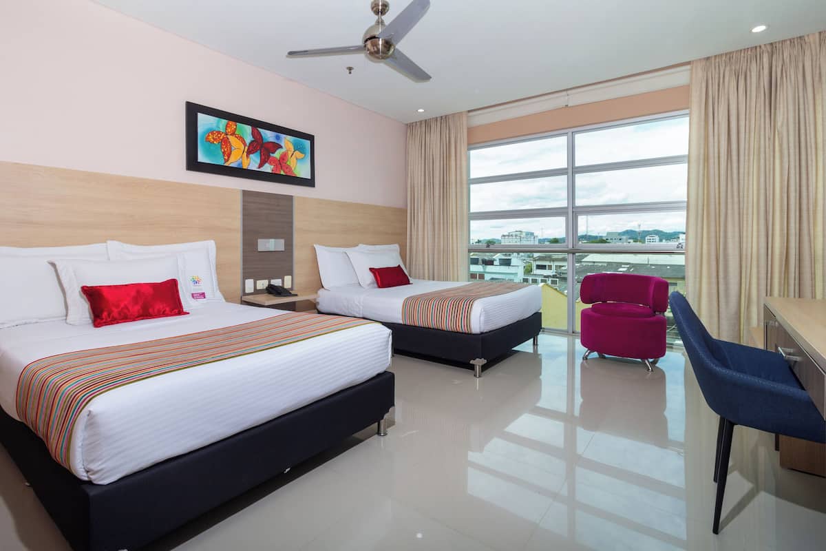 A hotel room with two beds, a desk, a chair, a ceiling fan, and a large window with a city view.