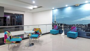 Un hall d’hôtel moderne doté d’une grande fenêtre donnant sur un paysage urbain, avec une rambarde en verre et des sièges aux couleurs vives.