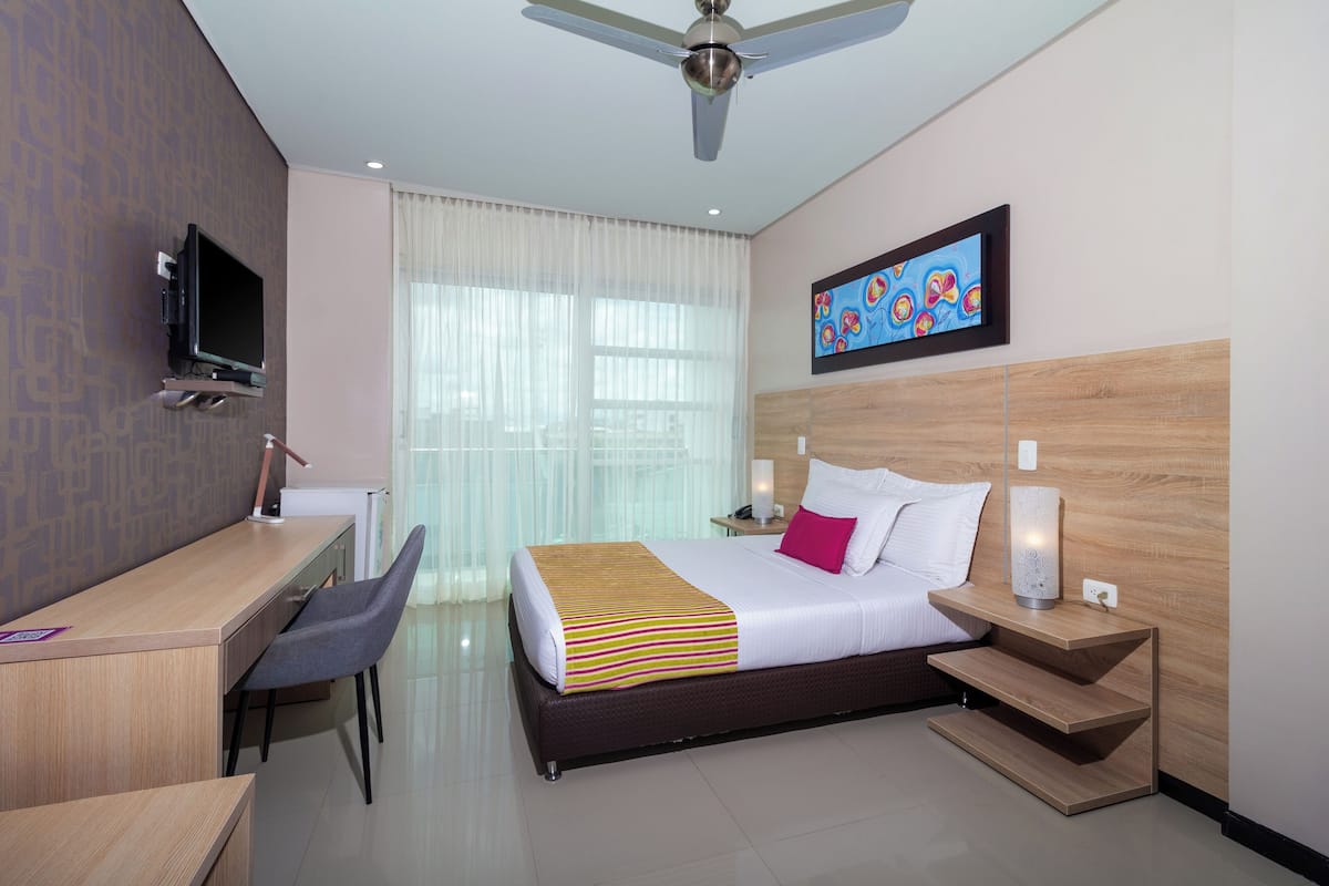 A hotel room with a bed, bedside tables, a desk, a chair, a television, and a ceiling fan.