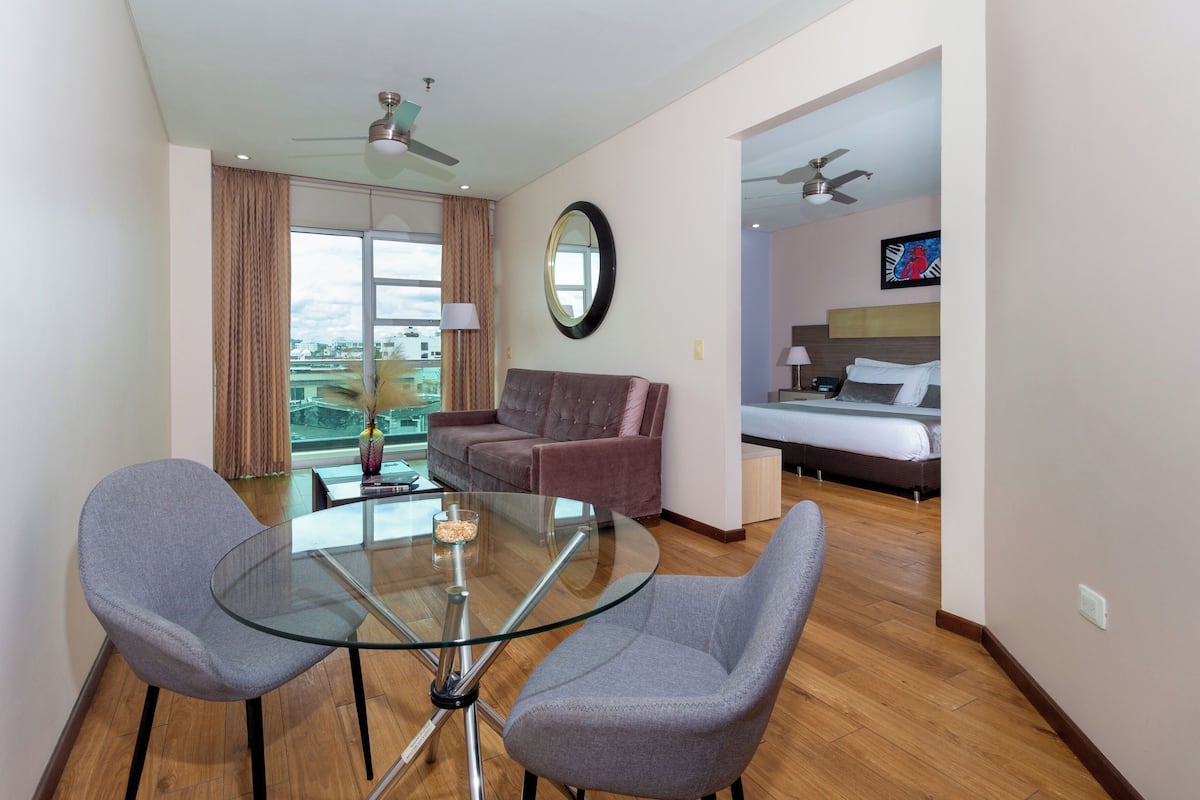 A hotel room with a glass dining table, two grey chairs, a sofa, a bed, a TV, and a balcony with curtains.