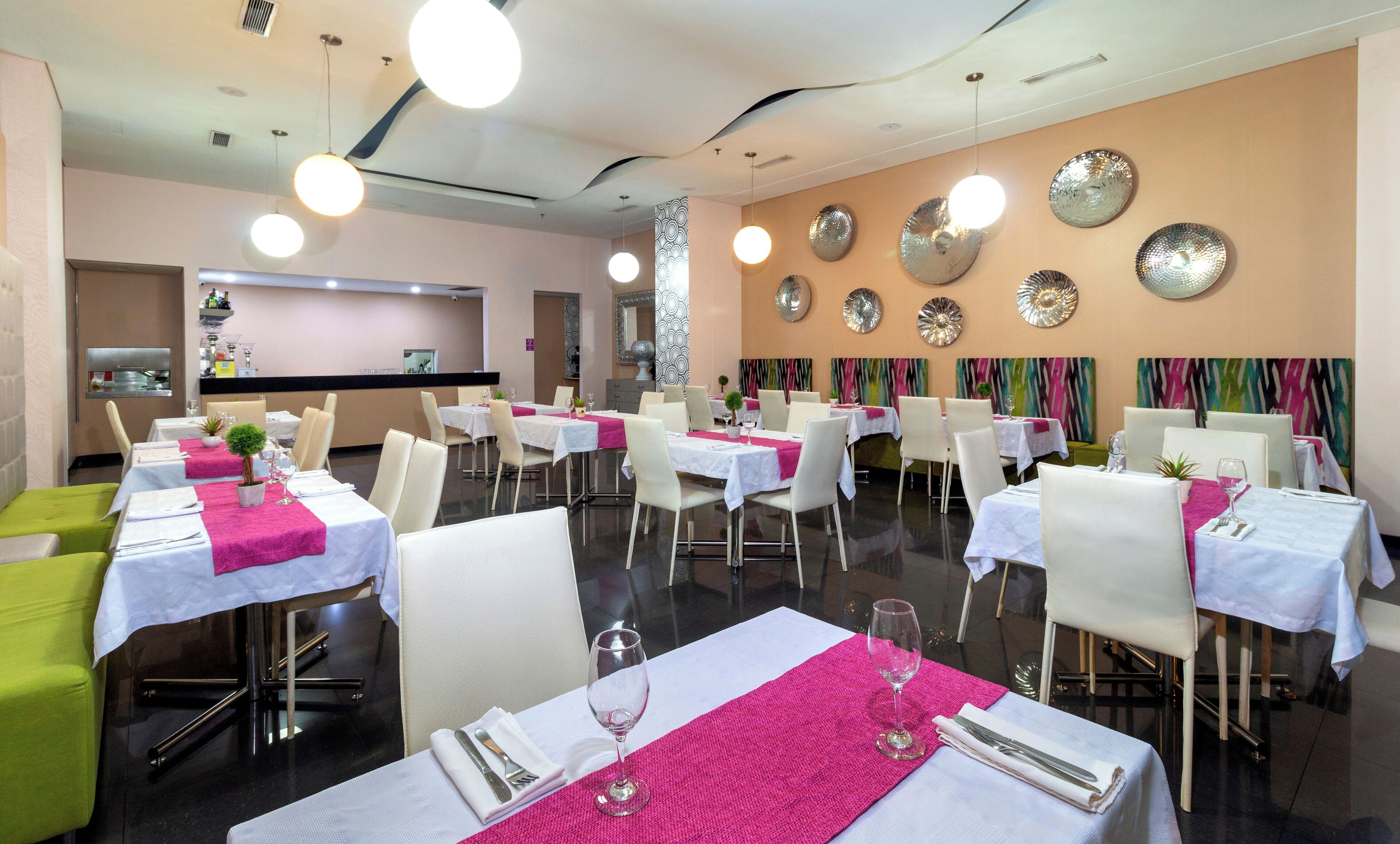 Een restaurant met meerdere tafels, elk voorzien van witte tafelkleden en roze placemats, met een modern interieur, abstracte muurkunst en hanglampen.