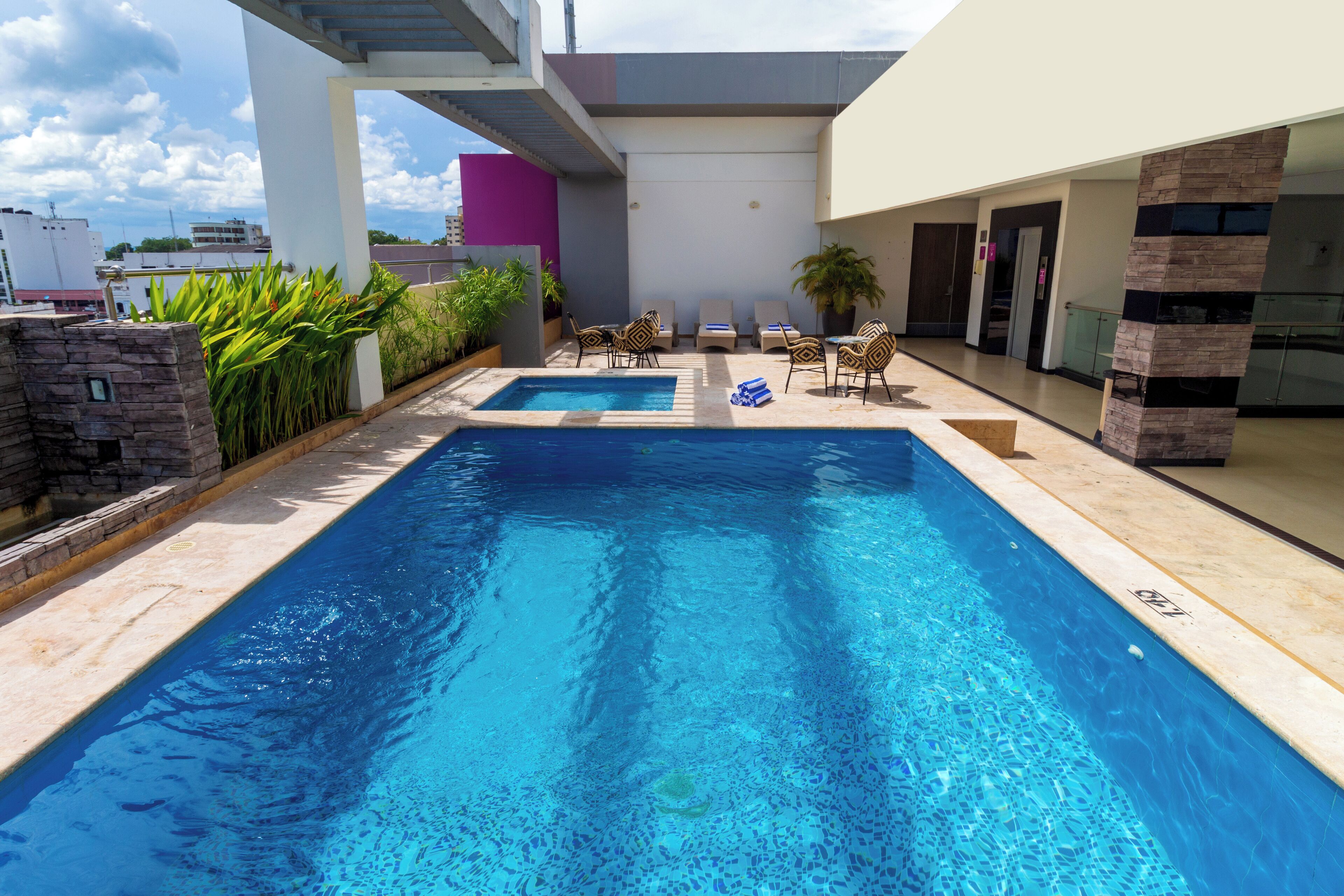 A swimming pool with two separate sections, surrounded by lounge chairs and potted plants, with a modern building in the background.