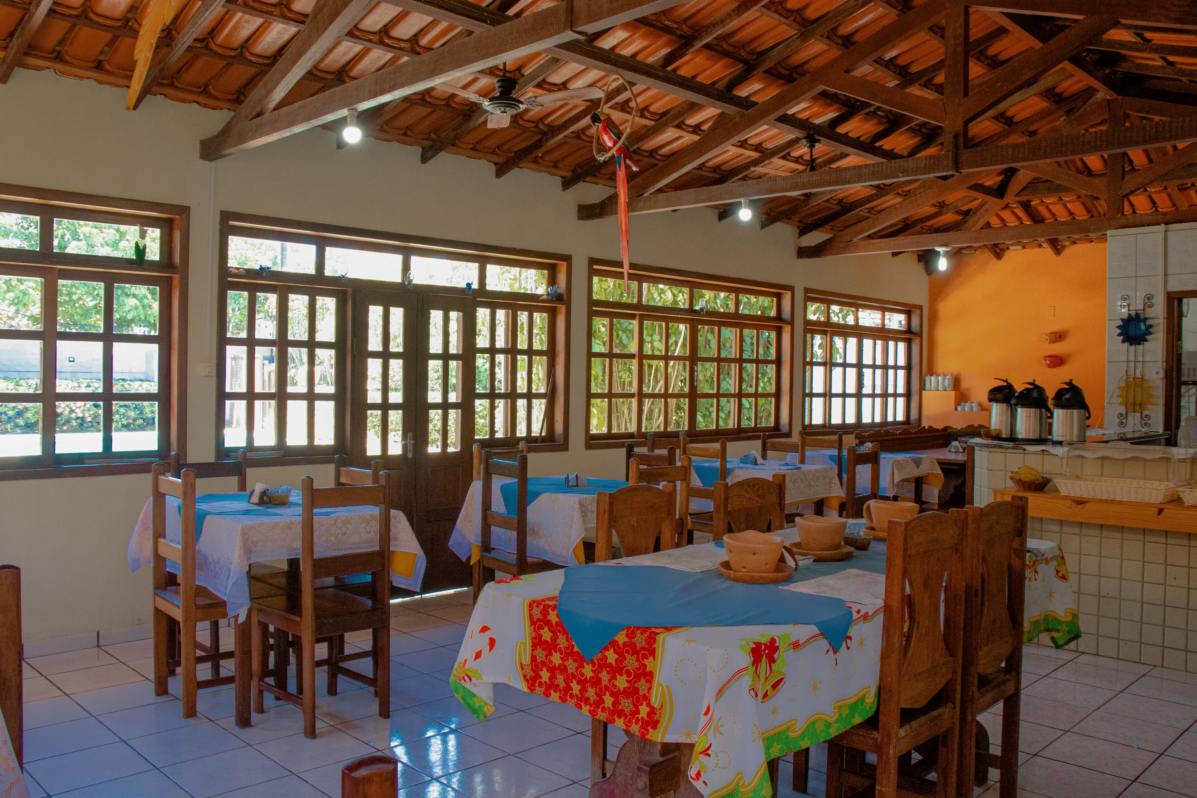 Un comedor con mesas y sillas de madera, ventanas amplias con marcos de madera y un suelo de baldosas.