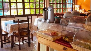 A breakfast buffet setup with wicker baskets containing fruits and pastries, a coffee machine, and a view of greenery through large windows.