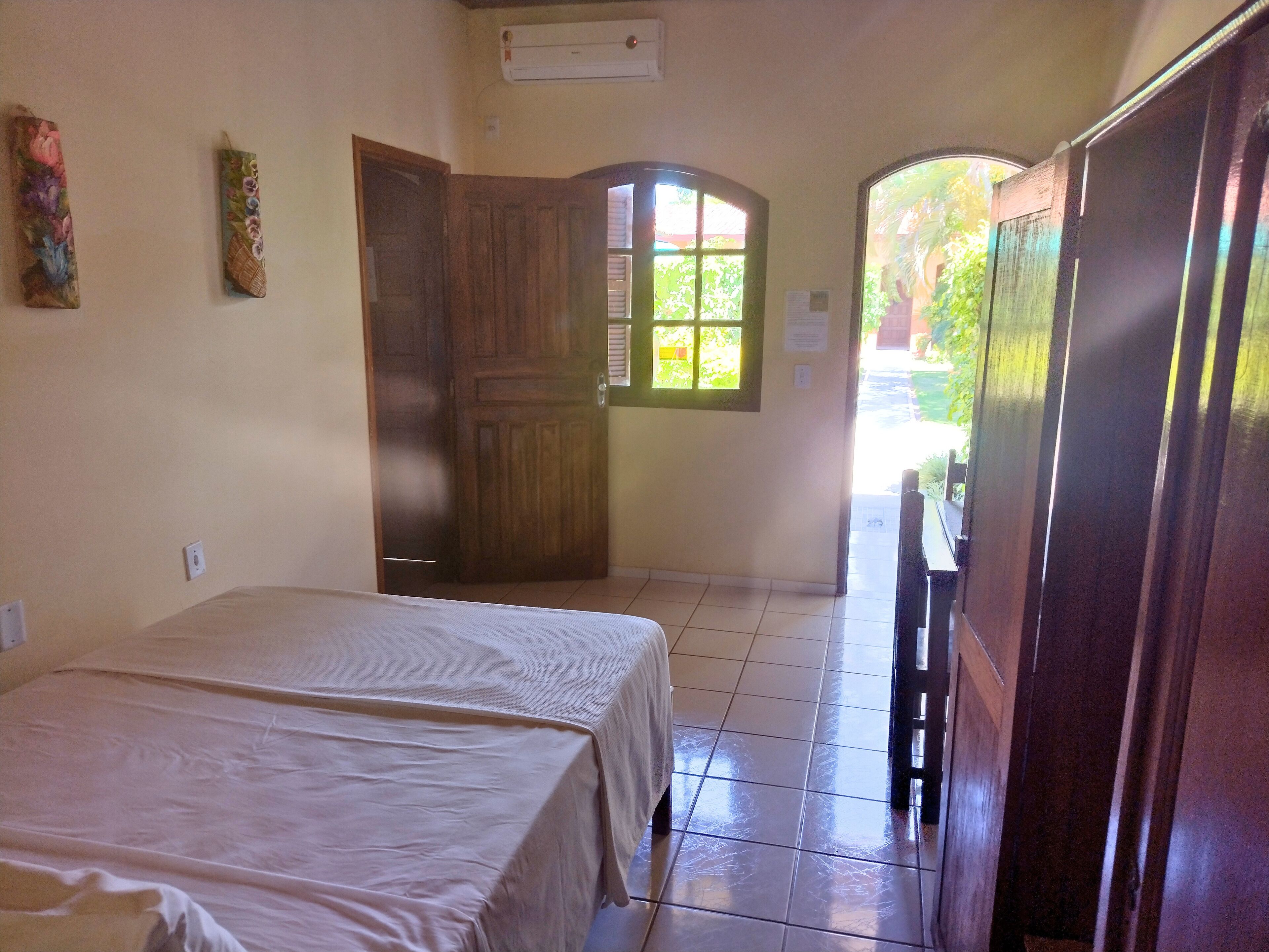 Une chambre d’hôtel avec deux lits, une armoire en bois, une porte donnant sur un balcon et une fenêtre avec vue sur la verdure.
