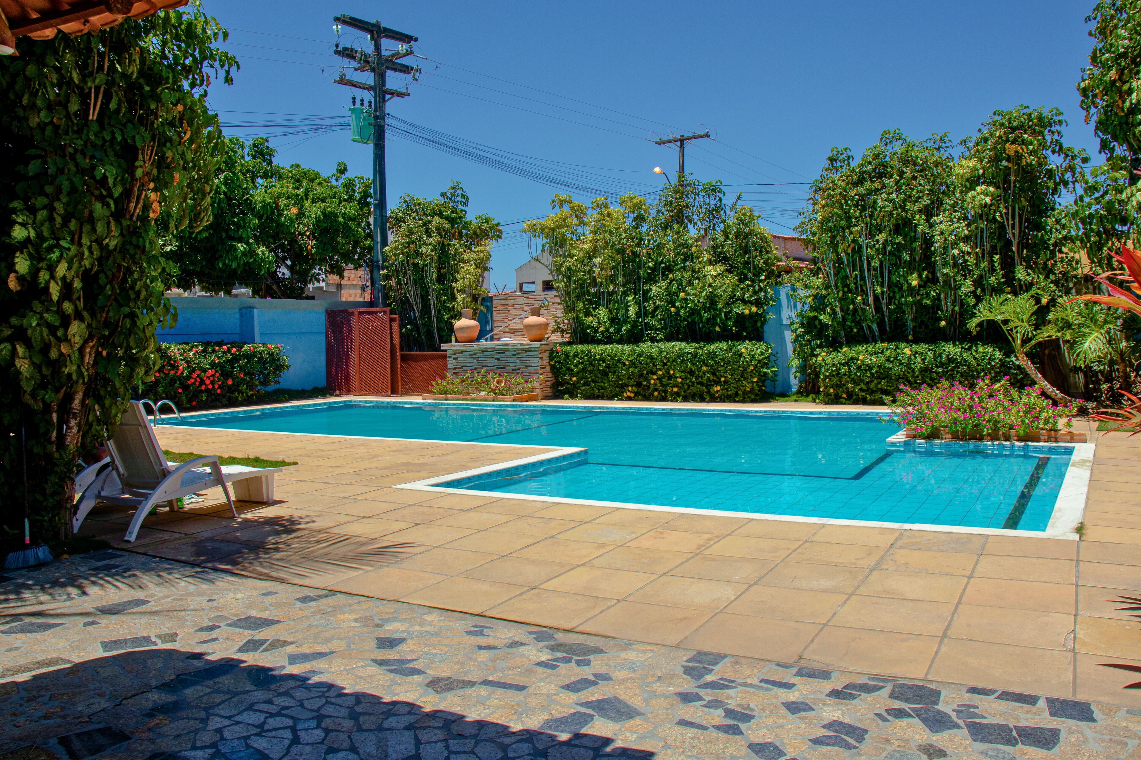 Una piscina rectangular con solárium de baldosas, rodeada de abundante vegetación y un muro de ladrillo.