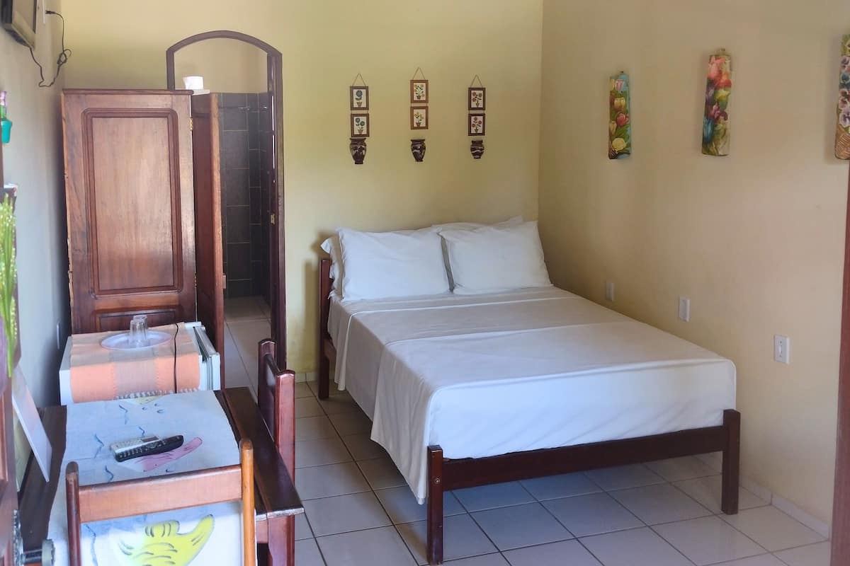 A bedroom with a wooden ceiling, a bed with white bedding, a wooden wardrobe, a desk with a chair, and a tiled floor.