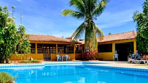 A swimming pool with a tiled deck, surrounded by palm trees and a yellow building with a tiled roof.