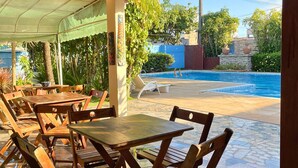 An outdoor patio with wooden tables and chairs, a pool, and a view of the surrounding garden and buildings.