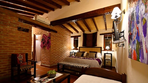 Una camera accogliente con un letto, una scrivania, una sedia e un tavolo con una pianta. Si vede una finestra con tende e una lampada a parete.