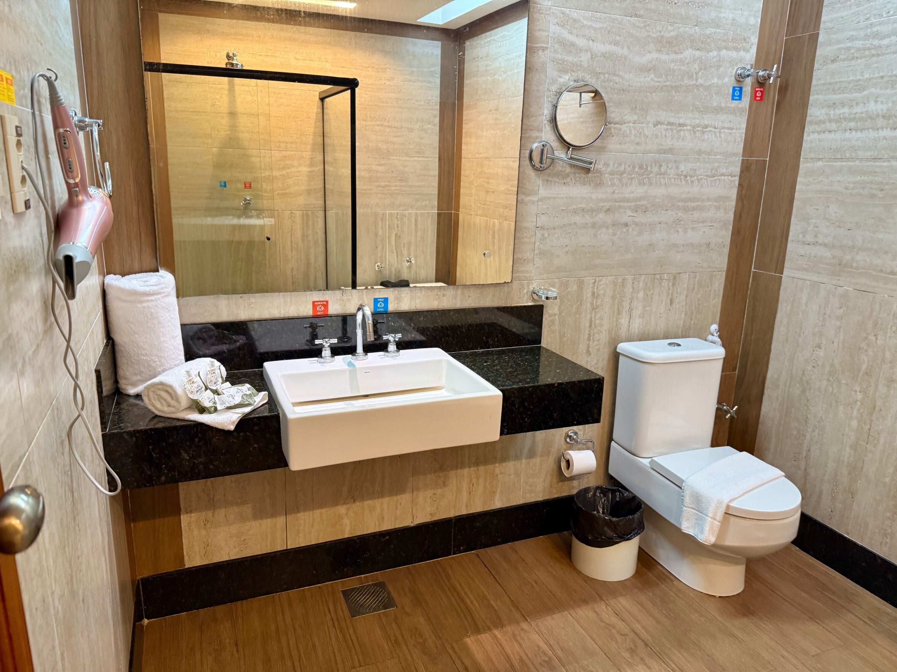 Un baño moderno con encimera negra, lavamanos blanco y inodoro. Hay una toalla y un cubo de basura en el suelo.