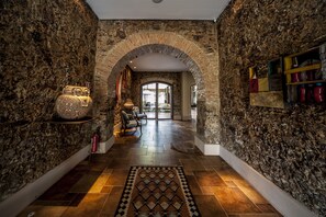 Un pasillo de paredes de piedra con un arco que da a una habitación. Hay una alfombra estampada en el suelo y una estantería con adornos en la pared de la izquierda.