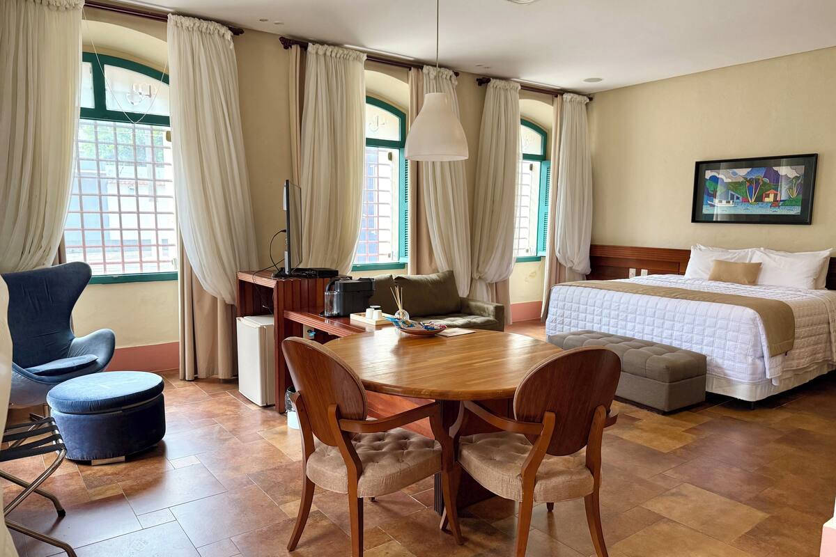 A hotel room with a wooden dining table, two chairs, a sofa, a bed with a white quilt, and a painting on the wall.