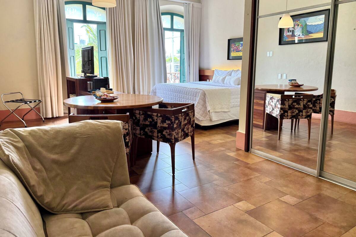 A hotel room with a bed, a dining table with chairs, a sofa, and a large mirror.
