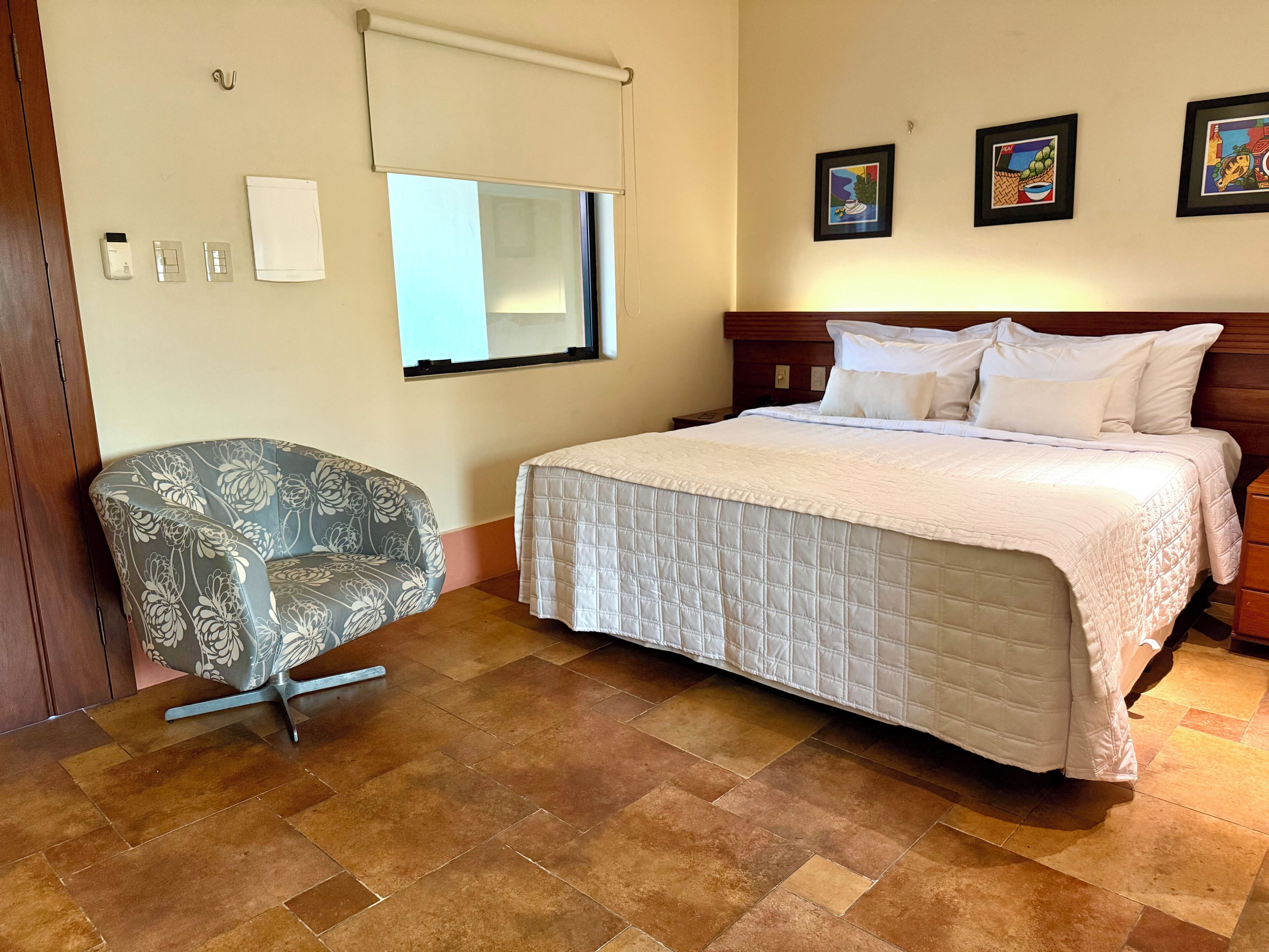 Habitación de hotel con cama, una silla, ventana con persianas, aire acondicionado instalado en la pared y cuadros enmarcados.