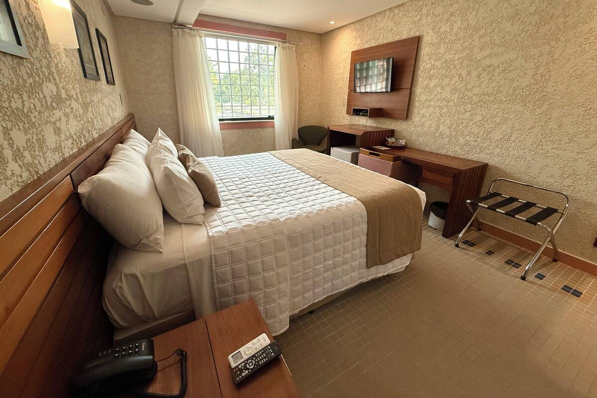 A hotel room with a bed, a desk, a chair, a TV, and a window with curtains.