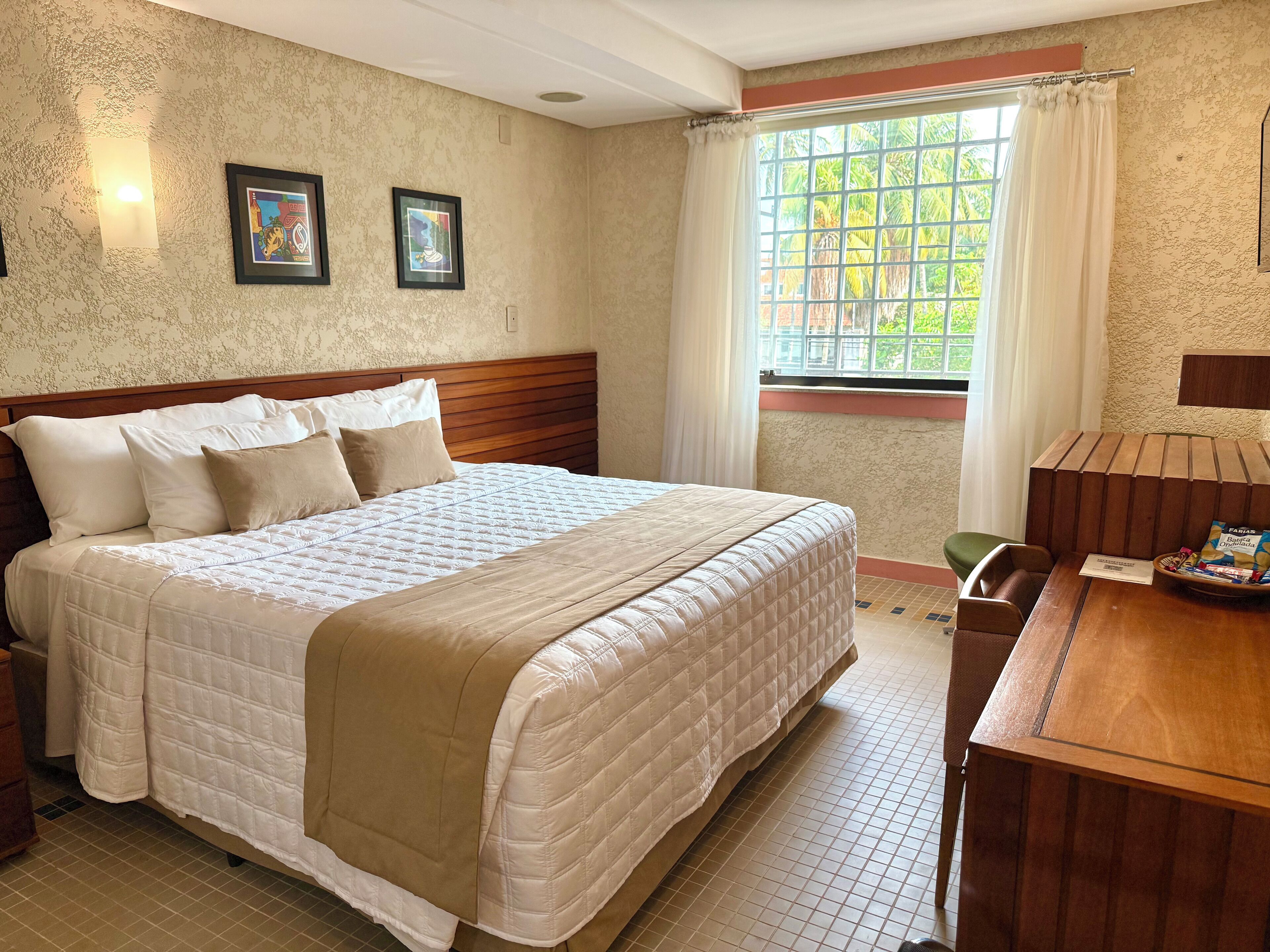 A hotel room with a large bed, a wooden headboard, a small table with a chair, and a window with curtains.