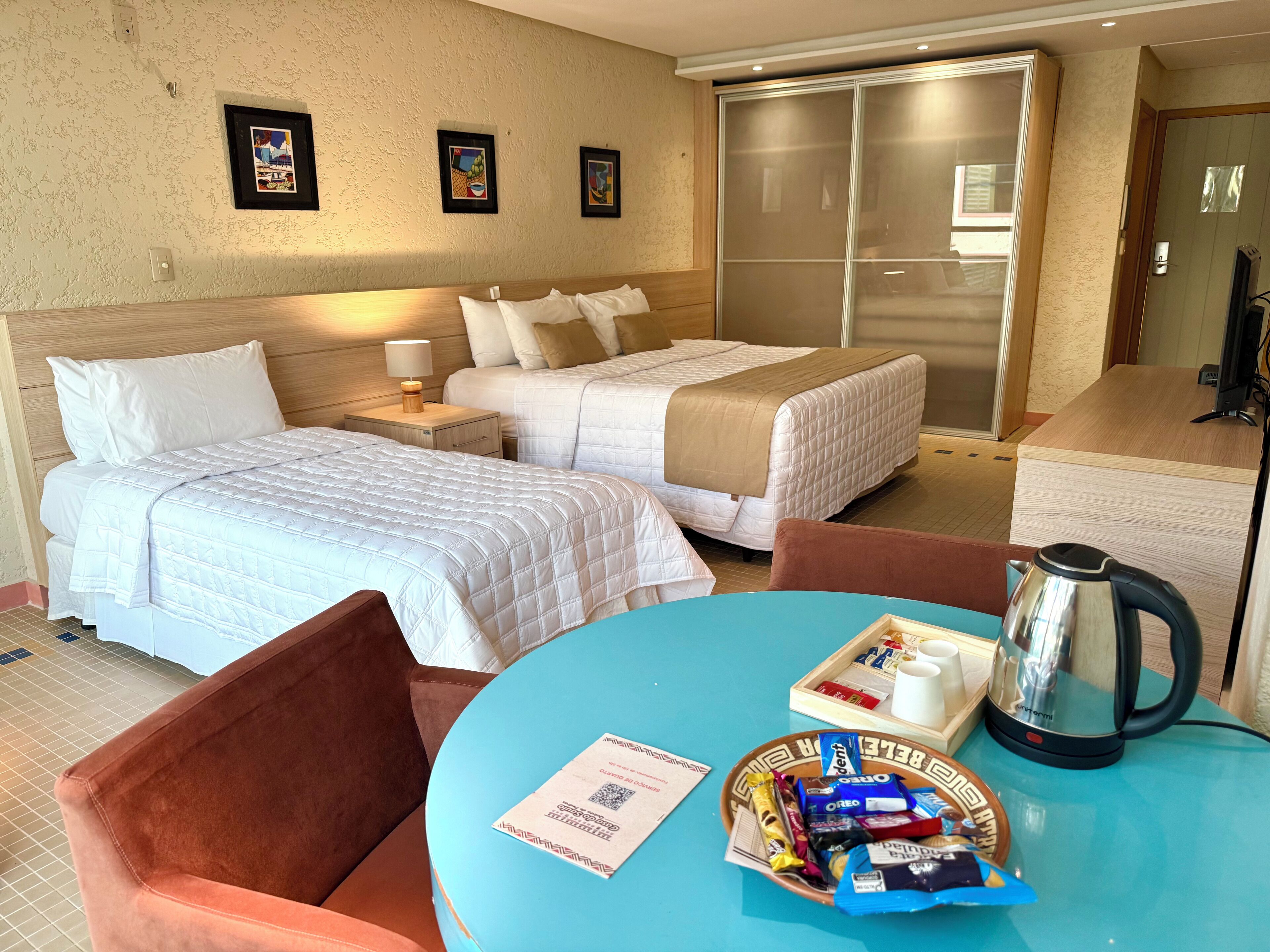 Quarto de hotel com duas camas, uma mesa de centro com petiscos, chaleira e armário com portas deslizantes.