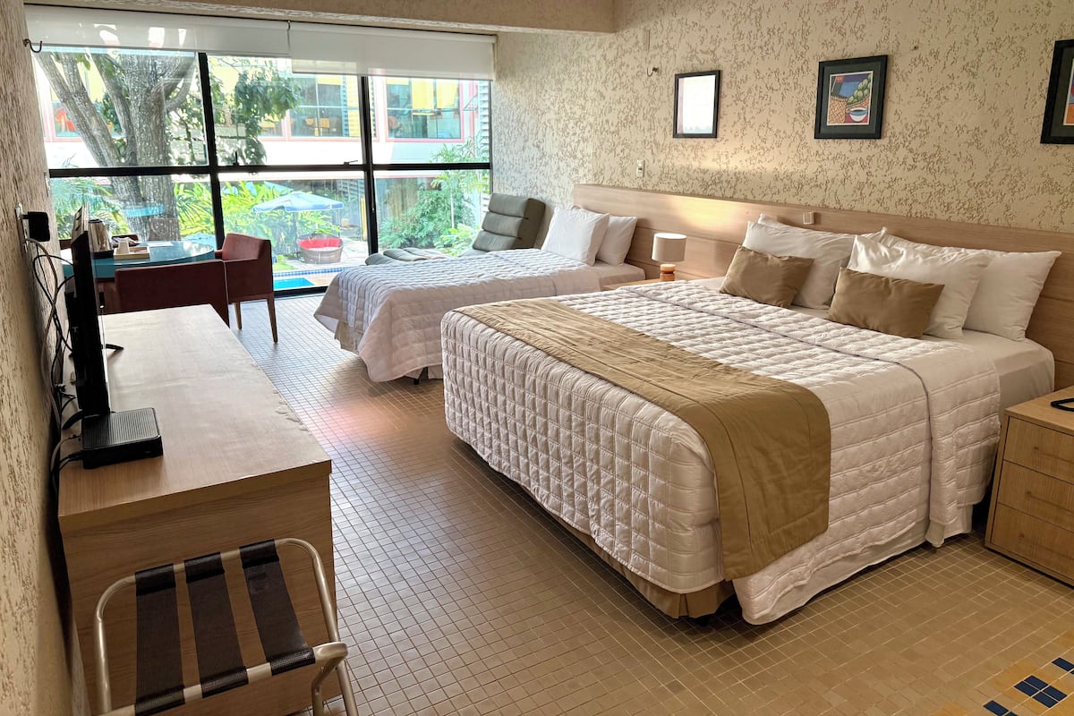 A hotel room with two beds, a desk, and a chair. There is a window with a view of the outside.